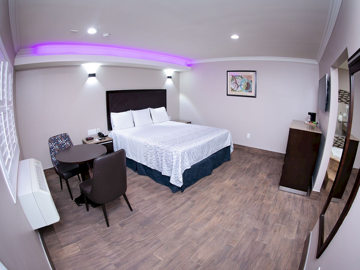 This image shows a modern hotel room with a large bed, a table with chairs, wall art, and a neon-lit ceiling, exuding a cozy ambiance.