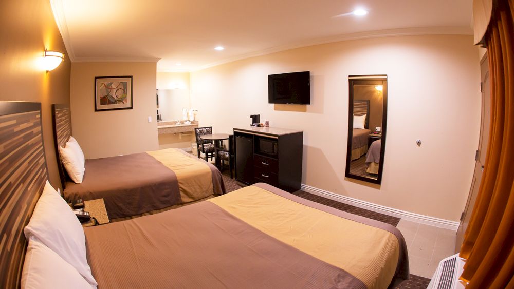 The image shows a hotel room with two beds, a TV, a mirror, a desk with a chair, a coffee maker, and a bathroom area in the background.
