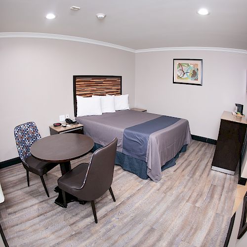 The image shows a modern hotel room with a bed, a table, two chairs, and a TV on the wall. The room has a light, neutral decor.