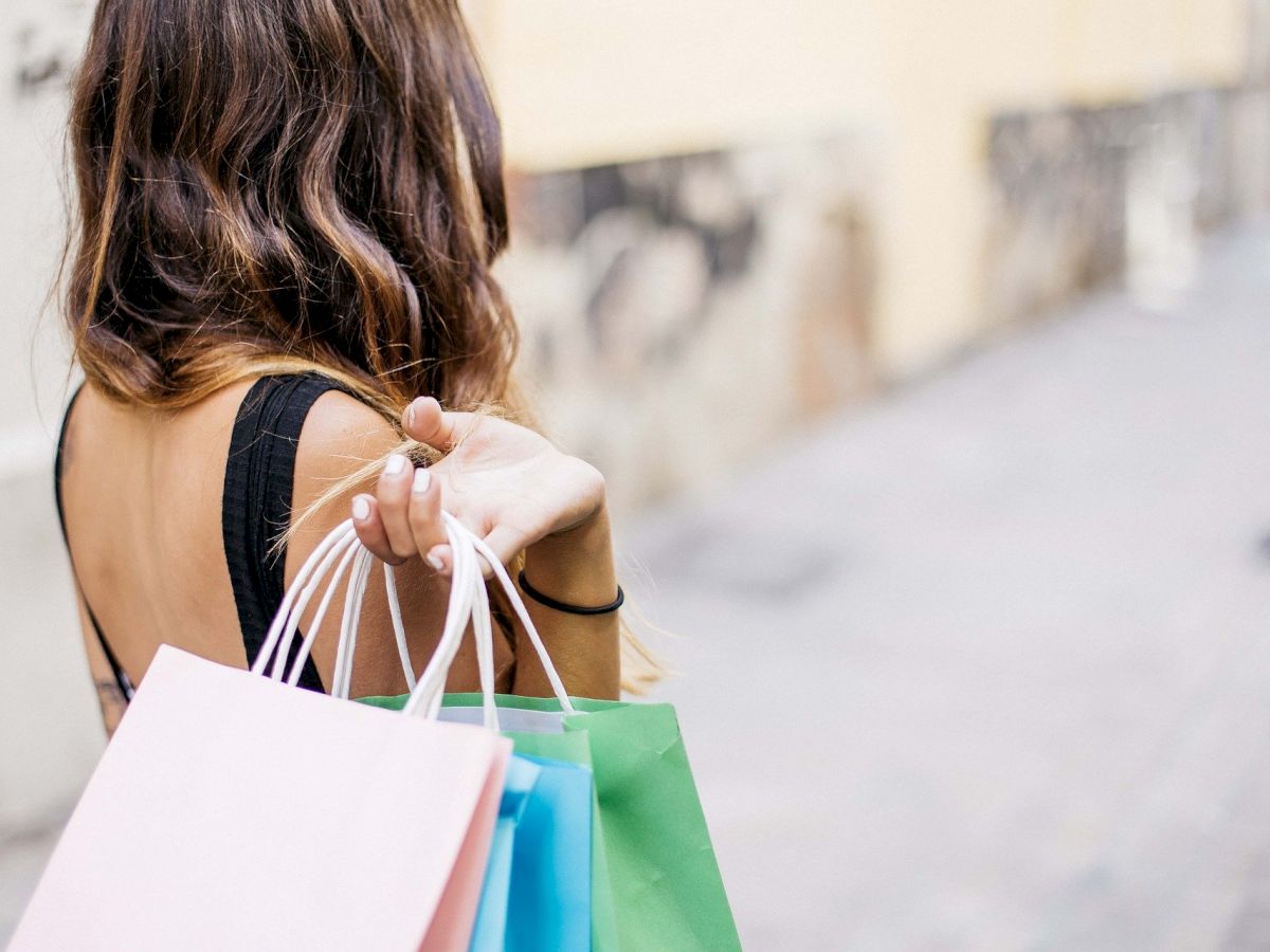 A person with long hair is walking down a street, holding several colorful shopping bags. The scene appears to be in an urban setting.