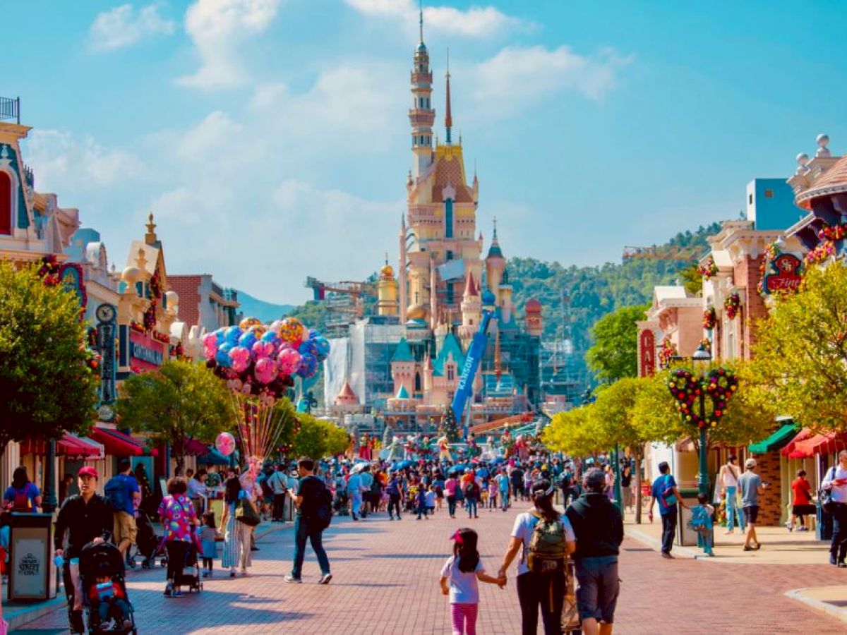 People are walking down a lively street towards a castle in a theme park, with balloons and colorful buildings around.