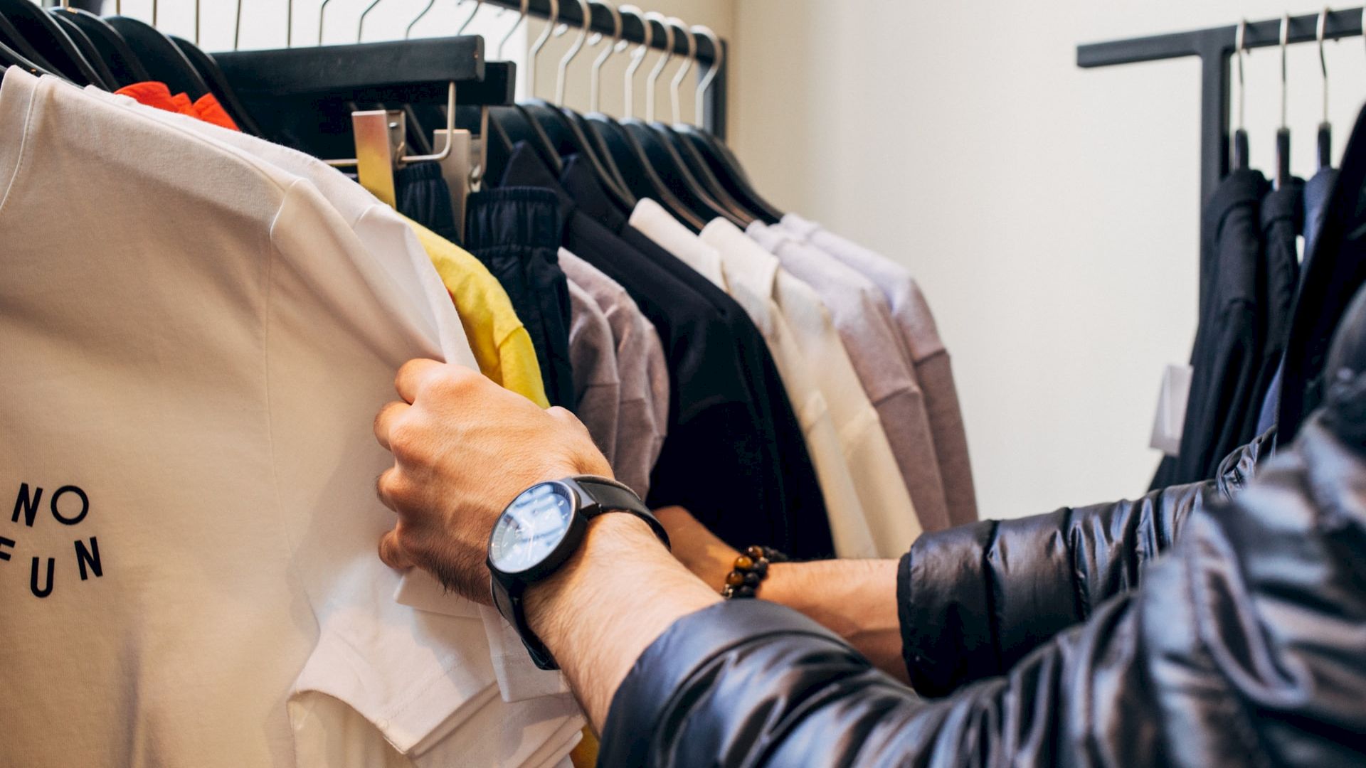 A person wearing a wristwatch is shopping for clothes, with their hands sifting through various colored shirts on a clothing rack.