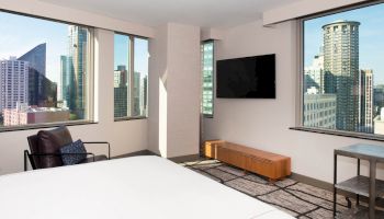 A modern hotel room with city views, featuring a bed, chair, wall-mounted TV, and minimalist furniture. Large windows show a skyline.