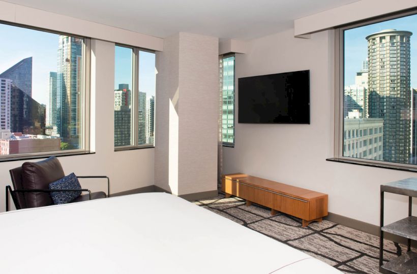 A modern hotel room with city views, featuring a bed, chair, wall-mounted TV, and minimalist furniture. Large windows show a skyline.