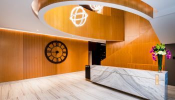 A modern reception area with wooden accents, a large clock on the wall, a marble counter, a spiral staircase, and decorative lighting and flowers.