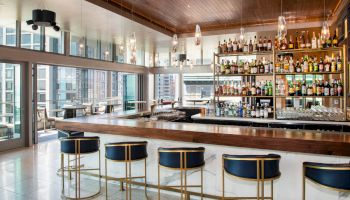 A sleek, modern bar with a long wooden counter, blue and gold bar stools, and a well-stocked liquor display, with city views visible through large windows.