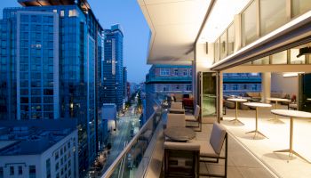 The image shows a modern cityscape with tall buildings and a stylish balcony at dusk, featuring tables, chairs, and an open sliding door area.