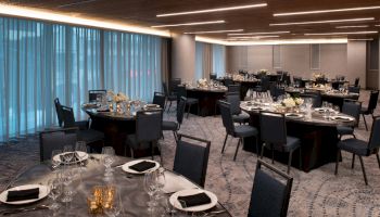 A modern event space with round tables set for dining, featuring black chairs, white flowers, and dim lighting, creating an elegant atmosphere.