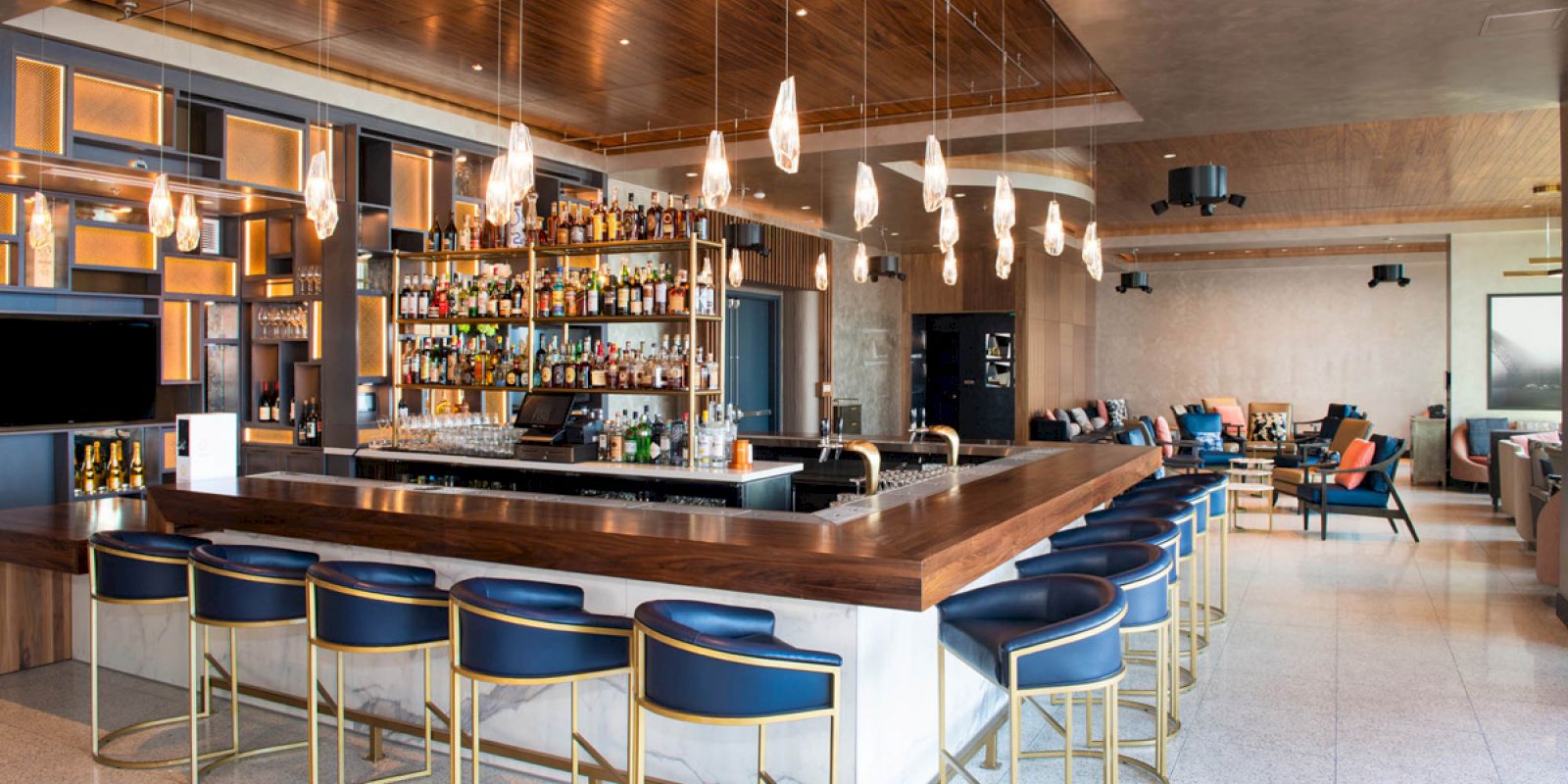 This image shows a modern bar with a wooden counter, blue bar stools, shelves of liquor bottles, pendant lights, and a lounge area in the background.