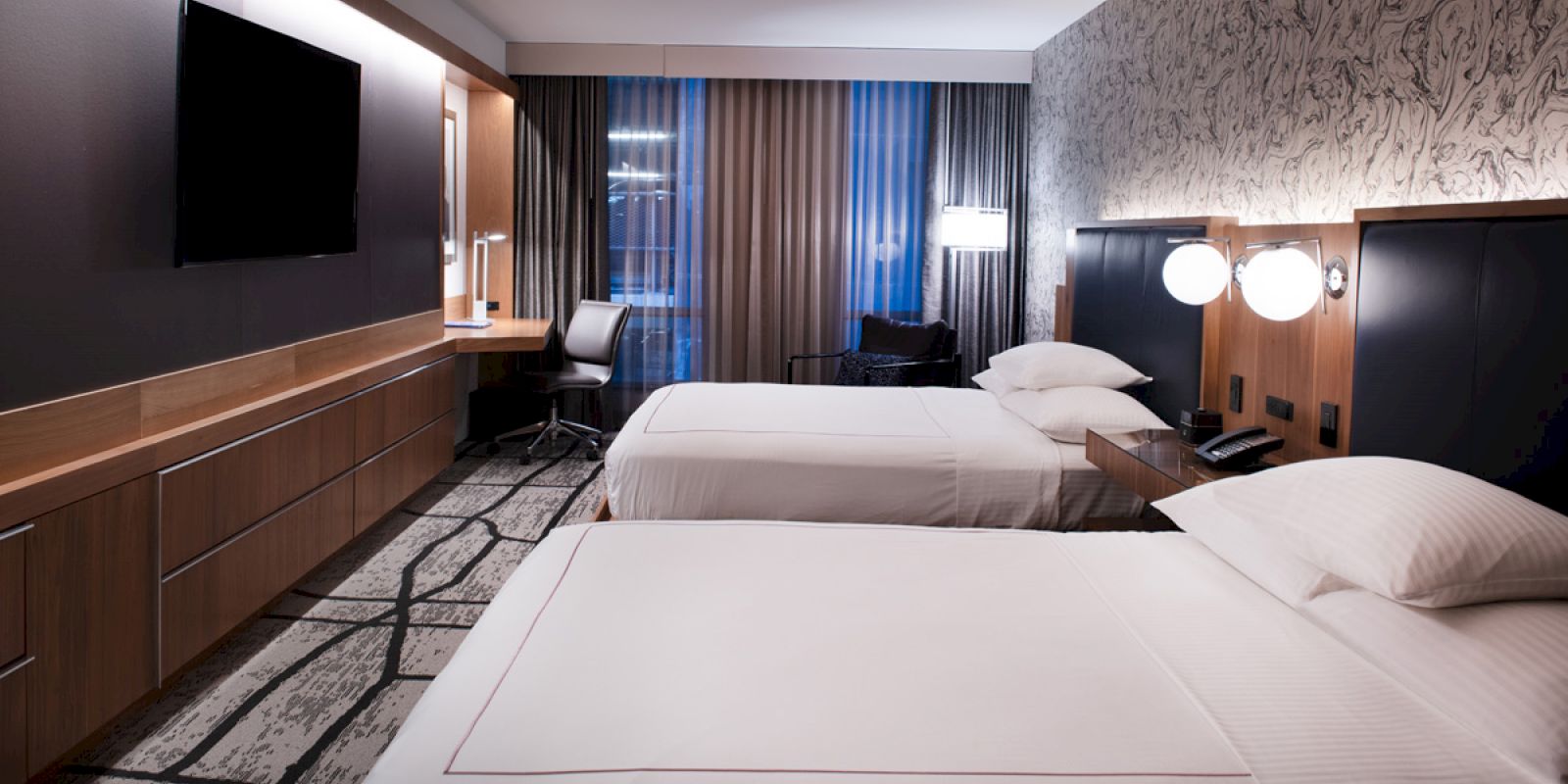 A modern hotel room with two neatly made beds, a wall-mounted TV, a desk, and a chair near the window, with stylish decor and lighting.