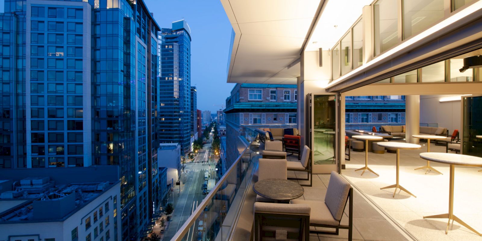 The image shows a modern, open balcony with seating, overlooking a cityscape with tall buildings and a street below, illuminated at dusk.