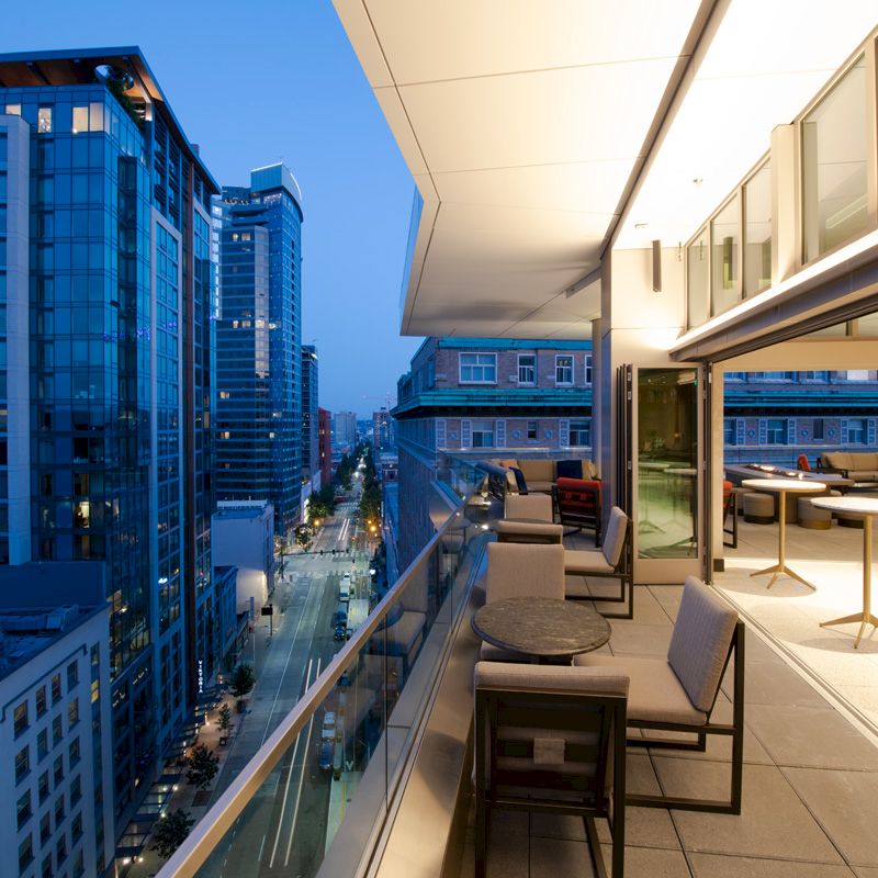 This image shows a modern cityscape with tall buildings at sunset, viewed from a balcony with seating and tables, featuring stylish outdoor furniture.