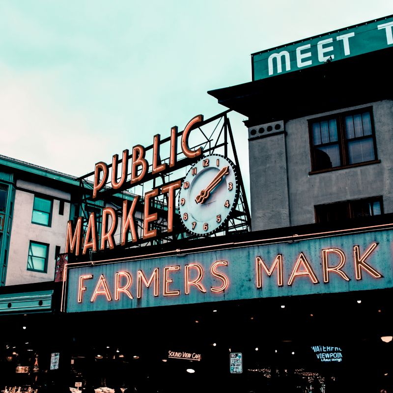 The image shows a well-known farmers market with a neon sign reading "PUBLIC MARKET" and "FARMERS MARKET," featuring a clock, in front of urban buildings.