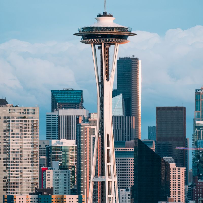 The image features the Space Needle in Seattle, with the city’s skyline and various high-rise buildings in the background, set against a cloudy sky.