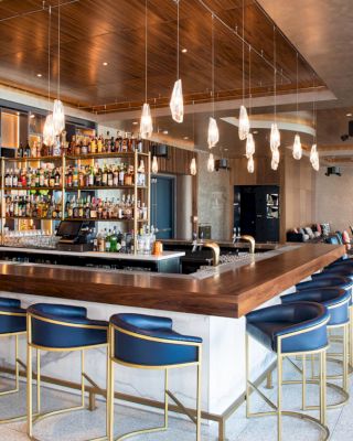 A modern bar with a wooden countertop, blue stools, hanging lights, and a wide selection of bottles on the shelves behind the counter.