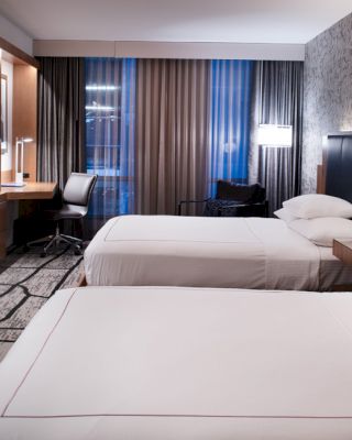 A modern hotel room with two neatly made beds, a desk with a chair, a wall-mounted TV, ambient lighting, and large curtains covering the windows.
