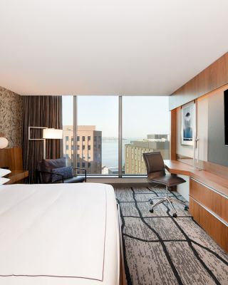 This image shows a modern hotel room with a large bed, desk, chair, and a flat-screen TV. A window provides a view of buildings outside.