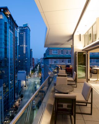 The image shows a view of a modern cityscape from a balcony at dusk, with buildings on the left and tables and chairs set up on the balcony.