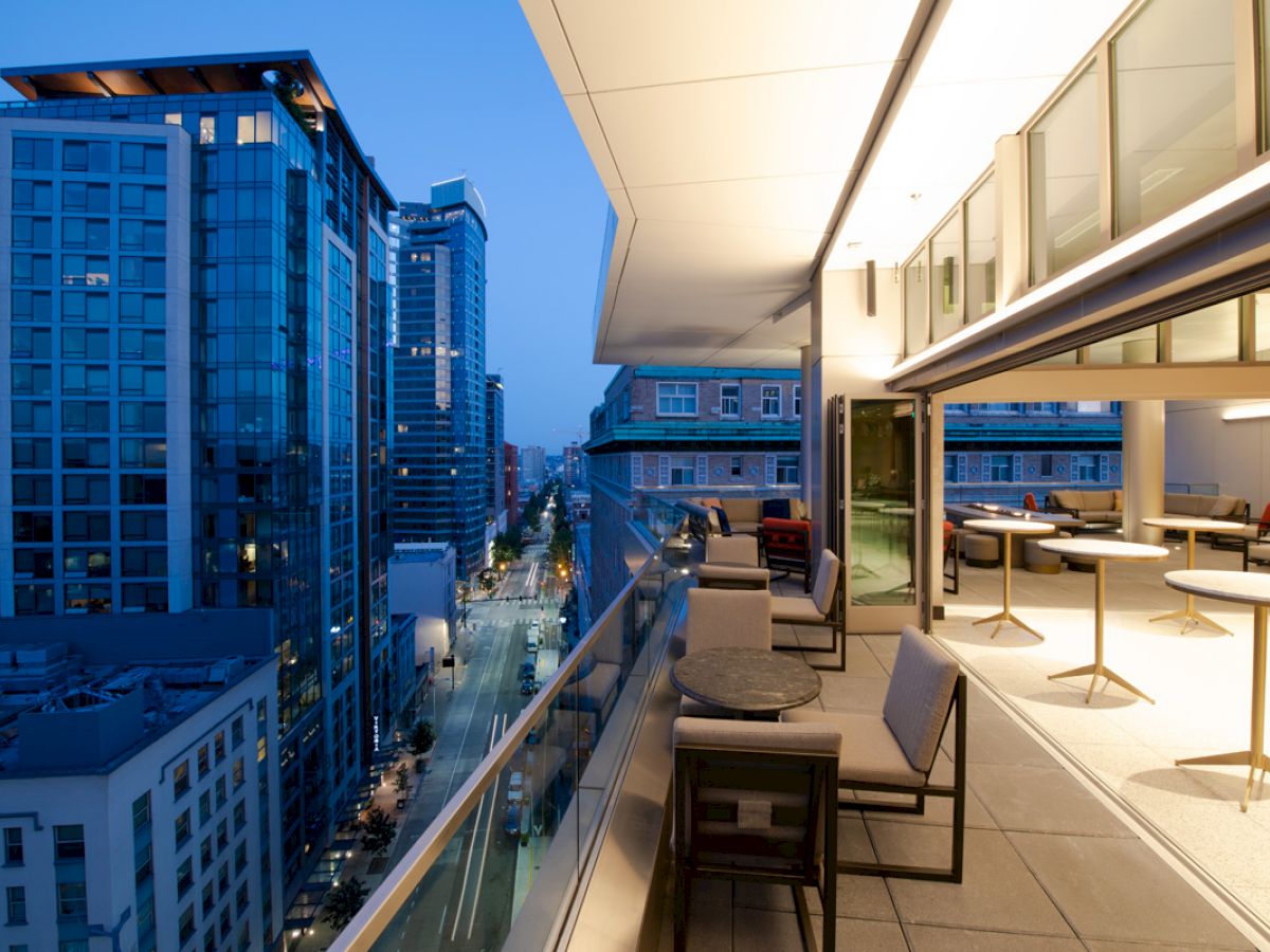The image shows a modern cityscape at dusk with high-rise buildings, viewed from a stylish balcony with seating and tables overlooking the street below.
