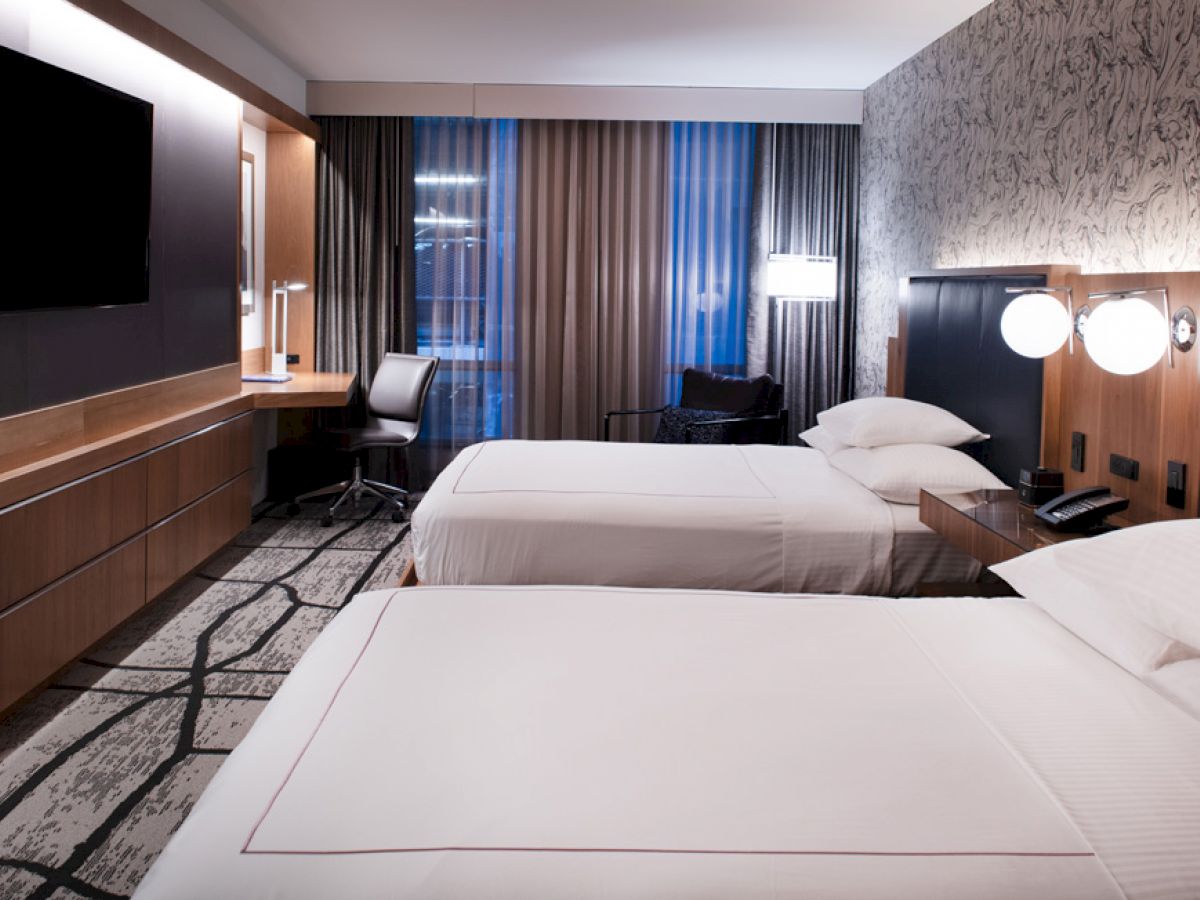 A modern hotel room features two neatly made beds, a wall-mounted TV, a desk with a chair, and a stylish decor with ample lighting and curtains.