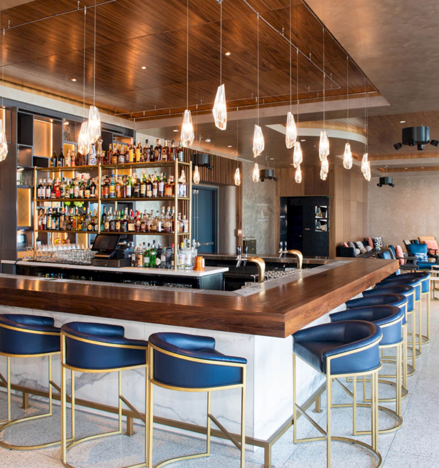 The image shows a modern bar with blue stools, a fully stocked bar shelf, hanging lights, and a stylish seating area in the background.