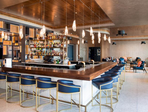 A modern bar with blue stools, pendant lights, and a wide selection of bottles on the shelves. There’s seating in the background and minimal decor.