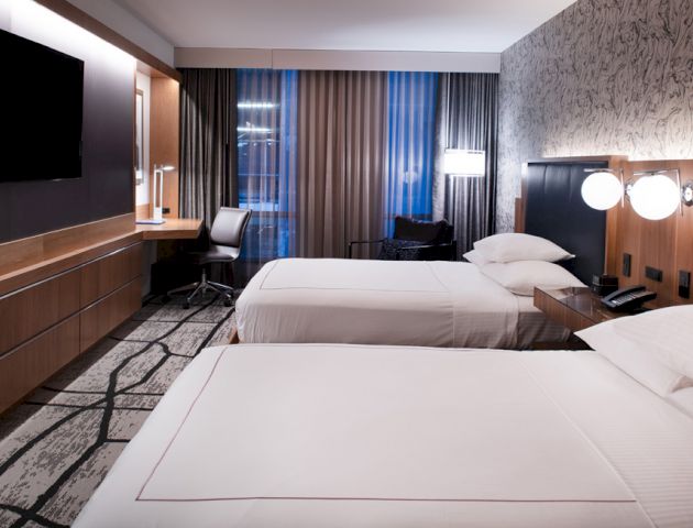 A modern hotel room with two double beds, a TV on the wall, a work desk, and large windows with curtains.