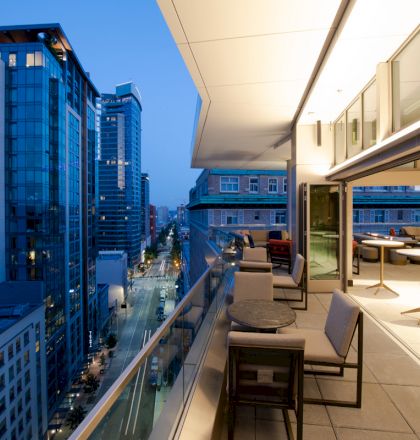 The image shows a modern cityscape with tall buildings and a balcony featuring outdoor seating and tables, overlooking a street at dusk.