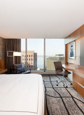 A modern hotel room featuring a large bed, a big TV mounted on the wall, a desk and chair, and a view of buildings and water through large windows.