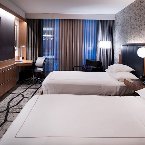 The image shows a modern hotel room with two beds, a flat-screen TV, a desk with a chair, and a large window with curtains, enhanced by wall lighting.