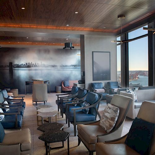 The image shows a modern lounge room with comfortable seating, a mural of a cityscape, large windows offering ocean views, and a Ferris wheel outside the windows.
