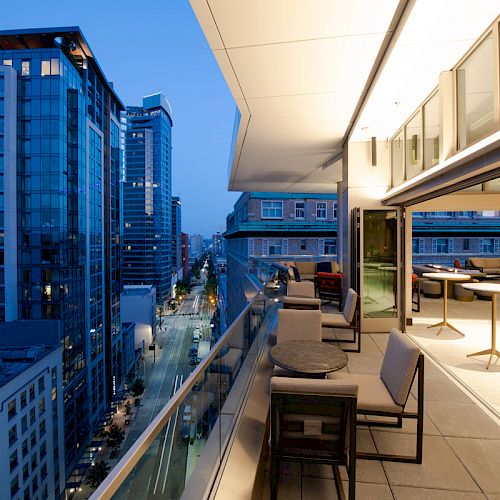 A modern balcony with outdoor furniture overlooks a cityscape filled with tall buildings and illuminated streets at dusk.