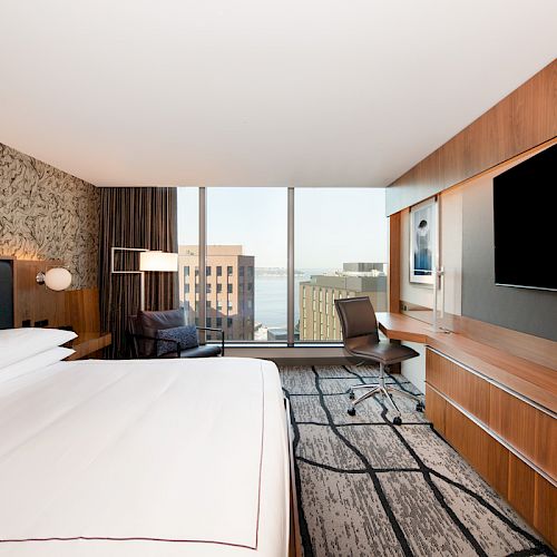 A modern hotel room featuring a large bed, a flat-screen TV, a desk with a chair, and a big window with a city view.