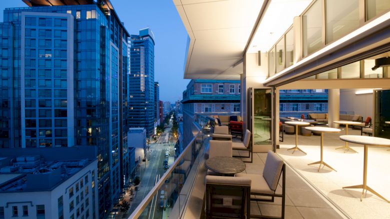 A modern cityscape in the evening featuring tall buildings and an upscale outdoor terrace with seating and tables.