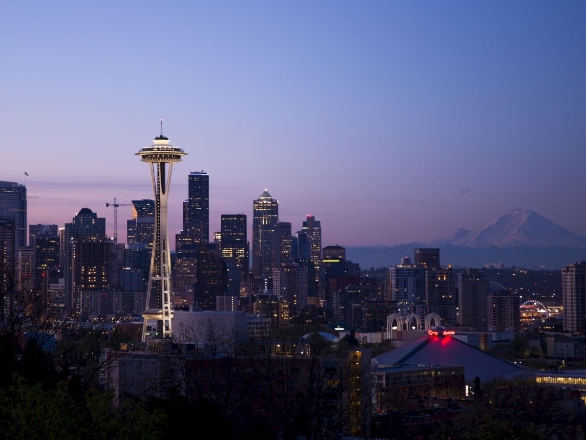 A cityscape view of Seattle at dusk, featuring the Space Needle prominently in the foreground and Mount Rainier visible in the background.