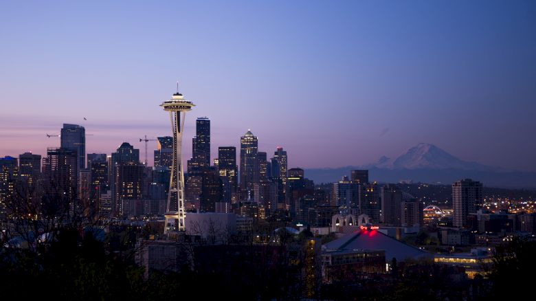 A cityscape view of Seattle at dusk, featuring the Space Needle prominently in the foreground and Mount Rainier visible in the background.