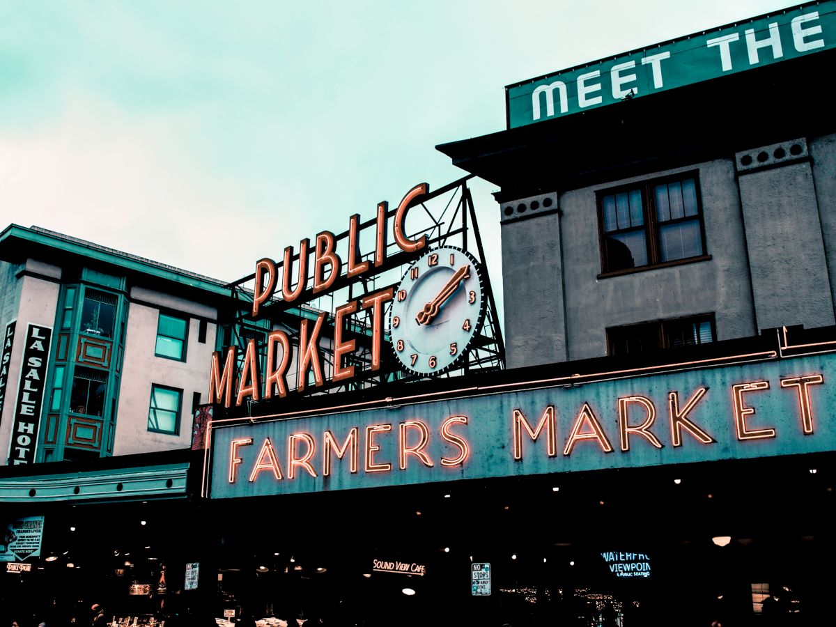 Image of a public market with neon signs reading "FARMERS MARKET." Buildings and a clock are also visible in the scene.
