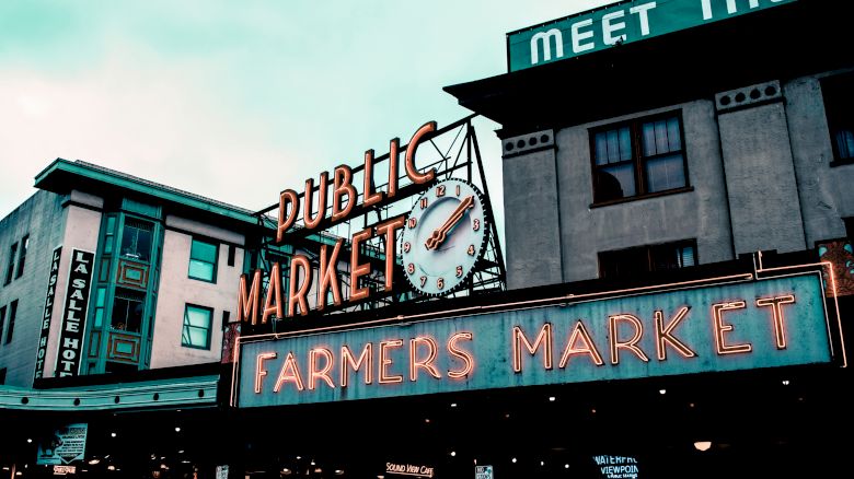 Image of a public market with neon signs reading "FARMERS MARKET." Buildings and a clock are also visible in the scene.