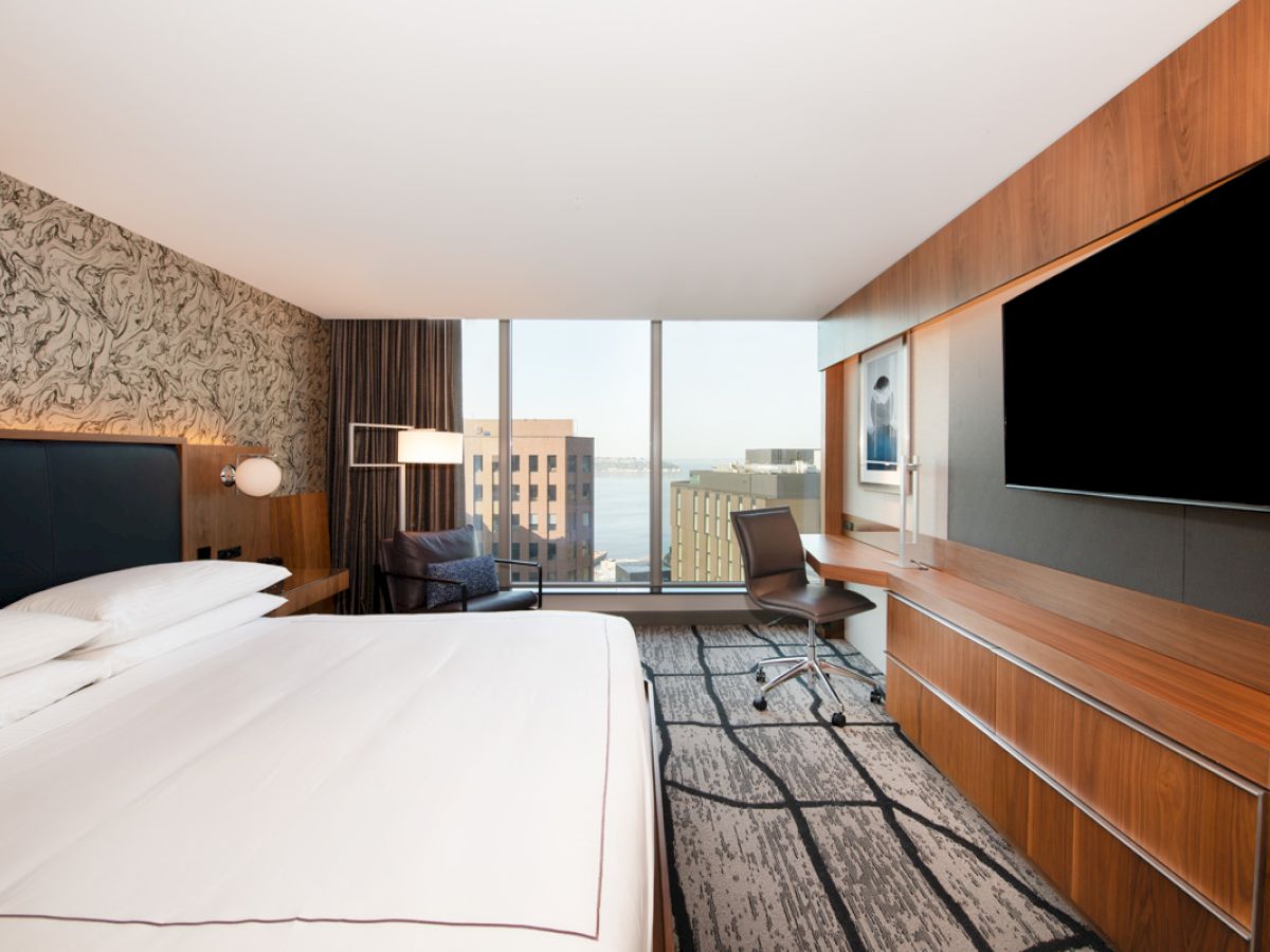 A modern hotel room features a large bed, a flat-screen TV, a desk with a chair, a patterned accent wall, and large windows overlooking a cityscape.