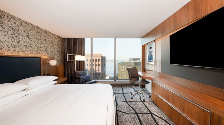 A modern hotel room features a large bed, a flat-screen TV, a desk with a chair, a patterned accent wall, and large windows overlooking a cityscape.