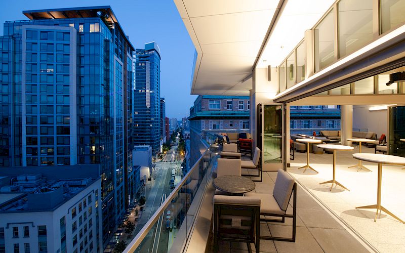 A modern outdoor patio with seating on a high-floor balcony overlooking a cityscape with tall buildings at dusk, illuminated by urban lights.