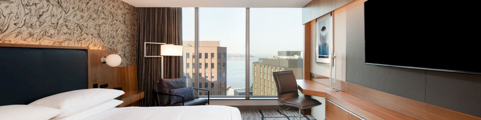 A modern hotel room features a large bed, TV, desk, chair, and a city view through large windows.