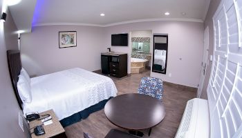 A hotel room with a bed, two chairs, a table, a dresser, a TV, and an adjoining bathroom. There is also an air conditioner by the window.