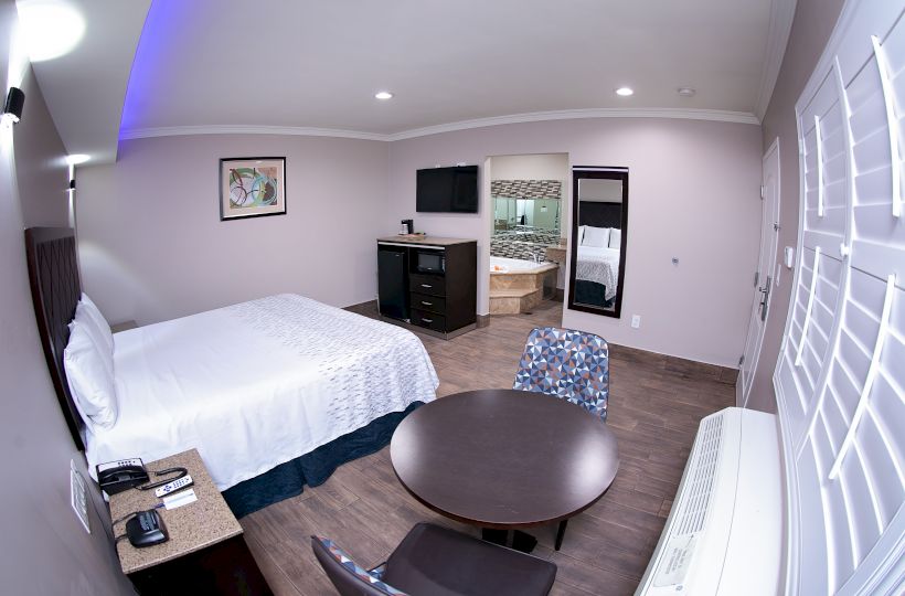 A hotel room with a bed, two chairs, a table, a dresser, a TV, and an adjoining bathroom. There is also an air conditioner by the window.