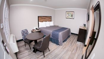 The image shows a modern hotel room with a queen bed, a small round table with two chairs, a dresser, and wall art.