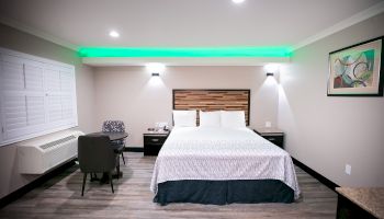 A modern bedroom features a large bed with wooden accents, a desk with a chair, a green light strip, a painting, and a light wood floor ending the sentence.