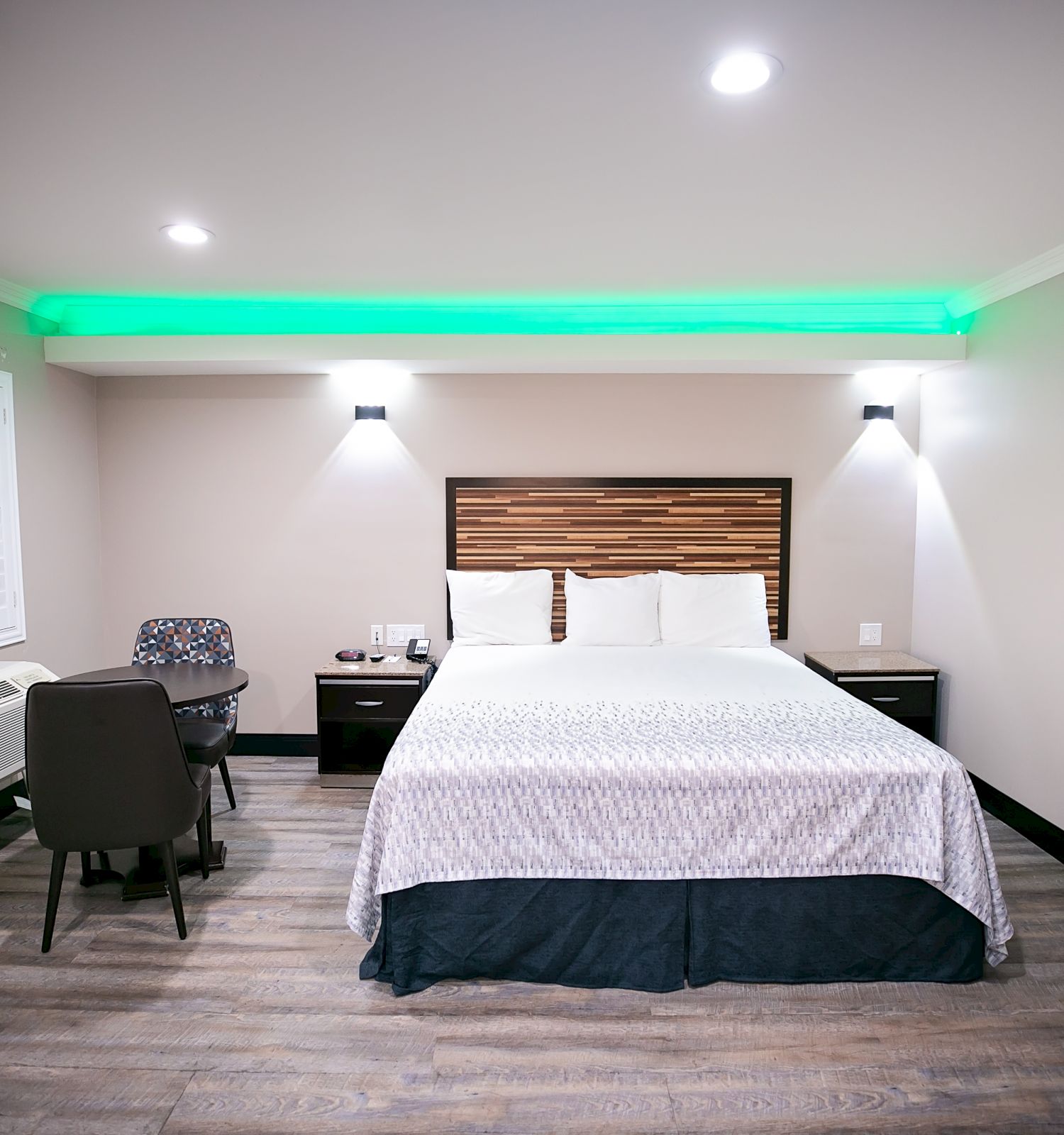 A modern hotel room with a king-sized bed, green ceiling lights, a work desk and chair, an air conditioner, and a framed abstract painting on the wall.