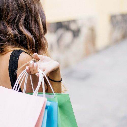 A person is walking down a street holding several colorful shopping bags in one hand. The person has long brown hair and is wearing a black top.