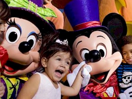 A young girl in a white dress poses joyfully with costumed characters resembling cartoon mice while other children and an adult enjoy the moment.